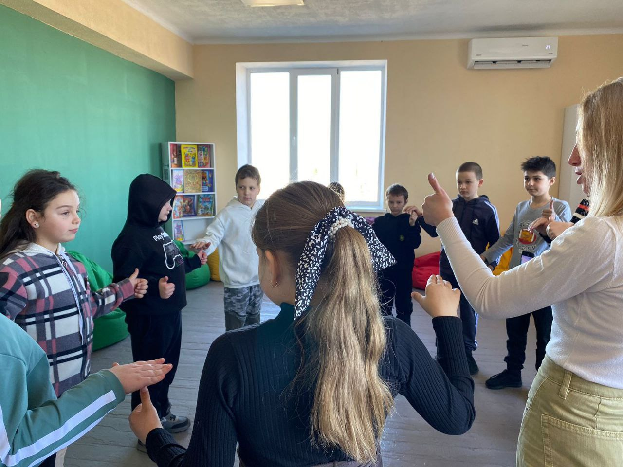 Група дітей та доросла жінка стоять у колі в світлій ігровій кімнаті. Вони виконують вправу на розвиток дрібної моторики або взаємодію, тримаючи руки перед собою та показуючи жести пальцями. На фоні видно яскраві крісла-мішки, полицю з книжками та велике вікно.