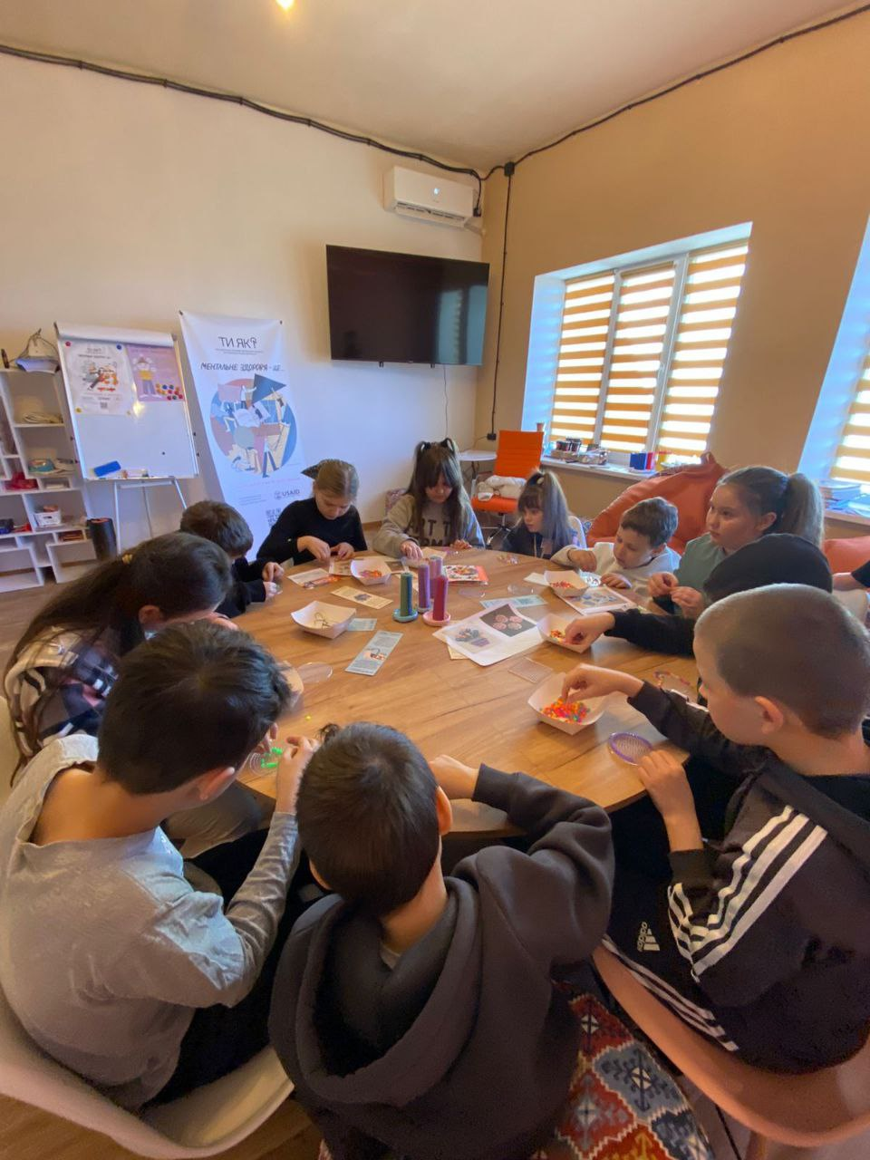 Діти за круглим столом займаються творчістю під час заходу програми ментального здоров'я «Ти як?». Вони працюють із дрібними деталями, ймовірно бісером або мозаїкою, розкладеними у контейнери. Позаду стоїть інформаційний стенд програми та облаштована зона відпочинку.