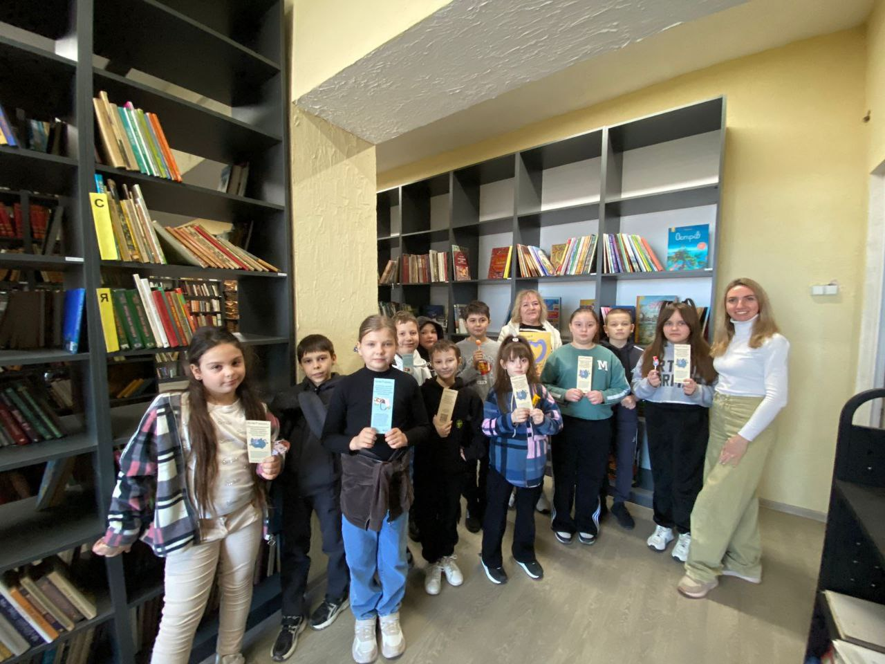 Група школярів та вчителька у бібліотеці. Діти тримають у руках саморобні книжкові закладки або листівки. На задньому плані — високі стелажі з книгами. У приміщенні панує дружня атмосфера освітнього заходу.
