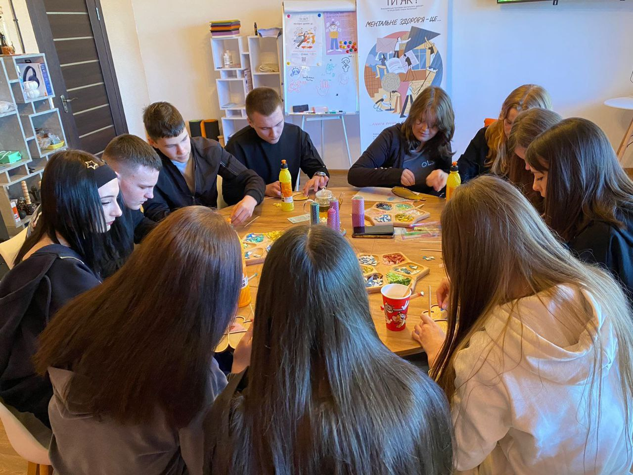 Група підлітків сидить за великим дерев'яним столом під час творчого заняття в межах програми «Ти як?». Учасники зосереджено працюють із дрібними деталями, ймовірно бісером або камінням, розкладеним у палітри. На фоні розташований інформаційний стенд про ментальне здоров'я.