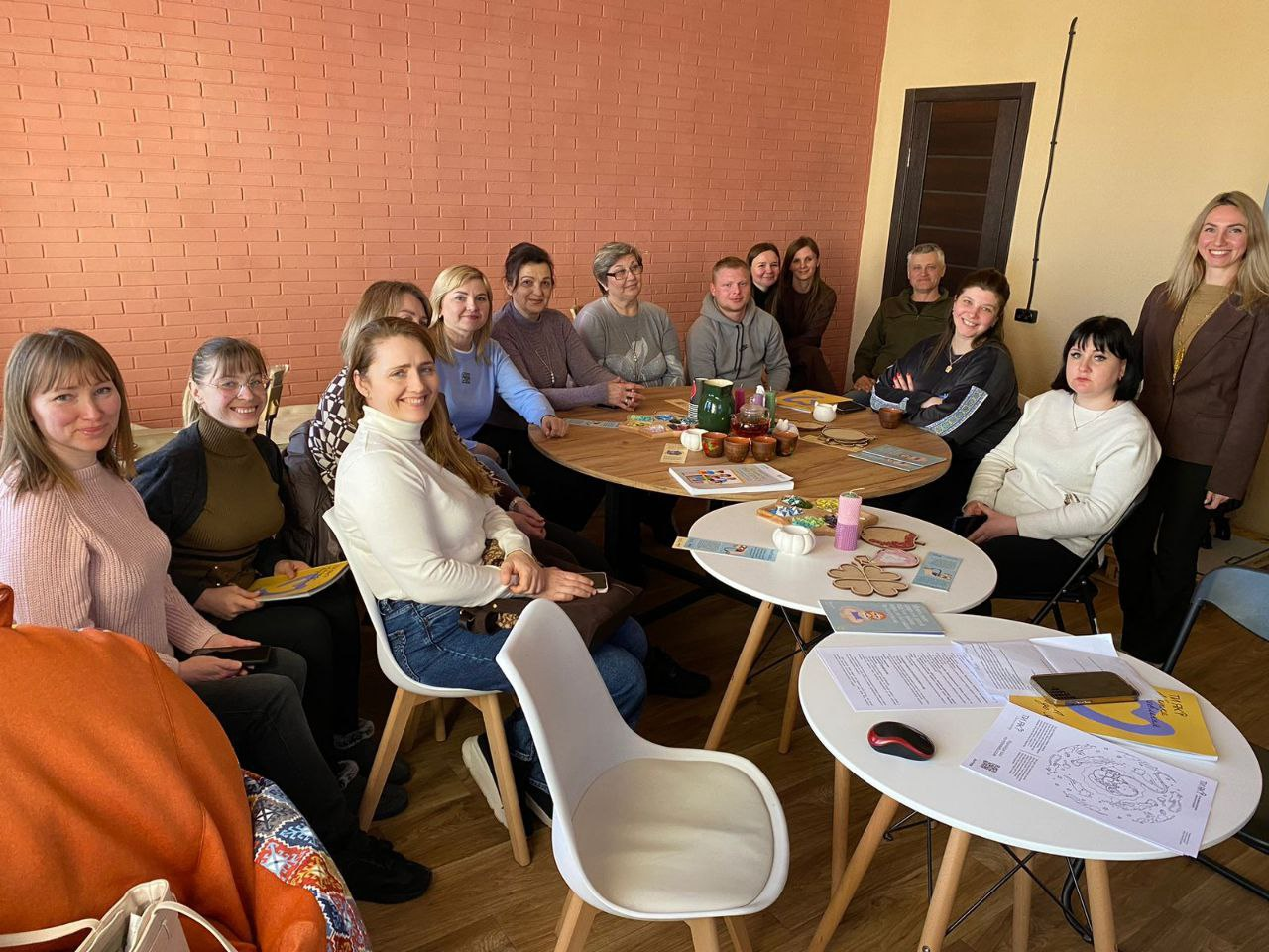 Група дорослих людей сидить навколо круглих столів під час психологічного тренінгу або групової терапії. На столах розкладені брошури програми «Ти як?», чашки для чаю та творчі матеріали. У приміщенні з цегляною стіною панує спокійна атмосфера діалогу.