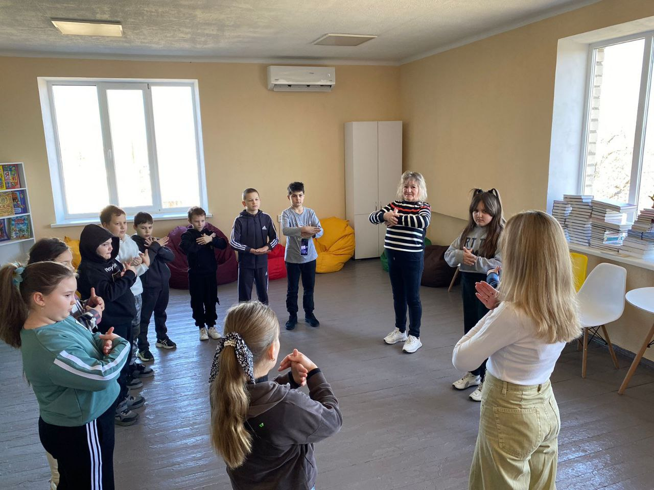 Група дітей та двоє дорослих стоять у колі в просторій світлій кімнаті. Вони зосереджено виконують вправу на психологічне розвантаження або координацію, повторюючи жести руками за інструкторкою. У приміщенні видно яскраві крісла-мішки, книжкові полиці та великі вікна.