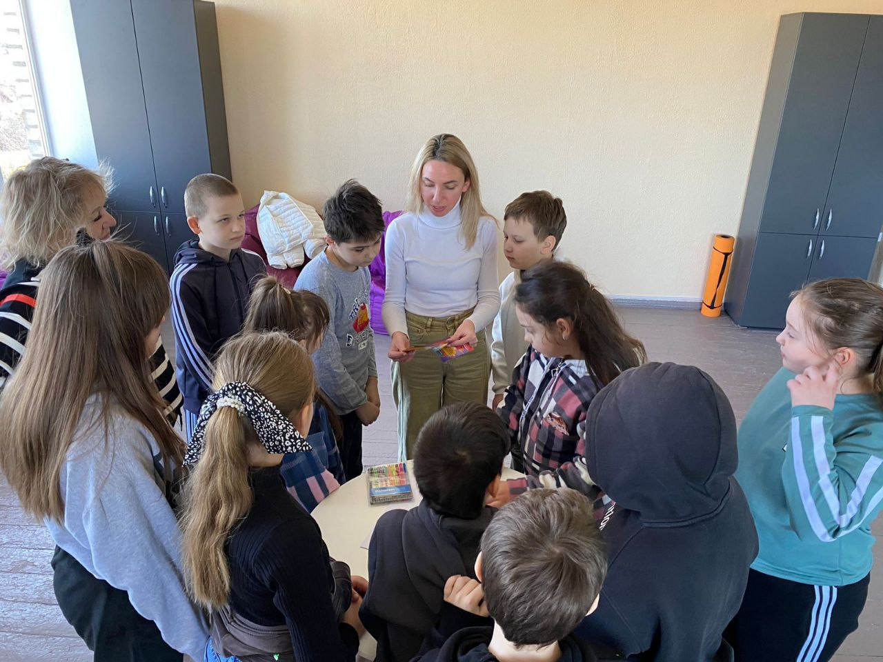 Група дітей обступила жінку-педагога біля круглого столу в світлій кімнаті. Доросла показує дітям маленьку кольорову картку або наклейку, а всі присутні уважно на неї дивляться. На фоні видно шафи, яскраві крісла-мішки та каремат.