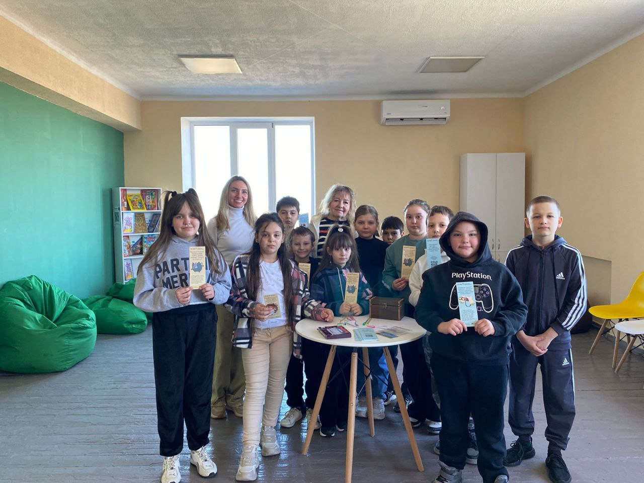 Група школярів та двоє дорослих стоять навколо круглого столу в сучасному освітньому просторі. Діти тримають у руках яскраві інформаційні буклети. У приміщенні з великим вікном видно зелені крісла-мішки, стелаж із книгами та світлі меблі.