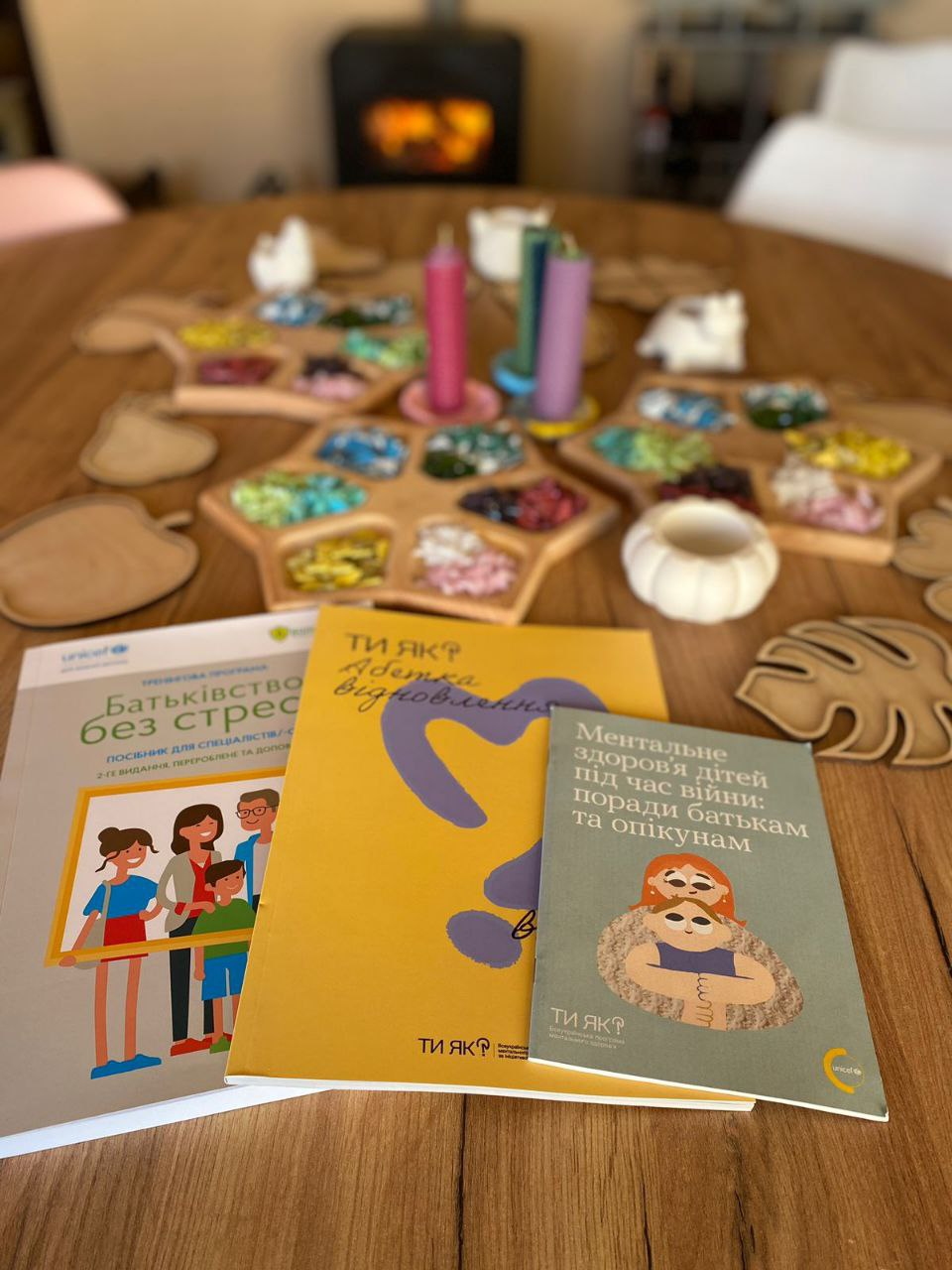 Три посібники з ментального здоров'я програми «Ти як?» та UNICEF лежать на дерев'яному столі. На передньому плані — книжки про батьківство без стресу та ментальне здоров'я дітей під час війни. На фоні — декоративне приладдя для арттерапії: палітри з камінням та кольорові свічки.