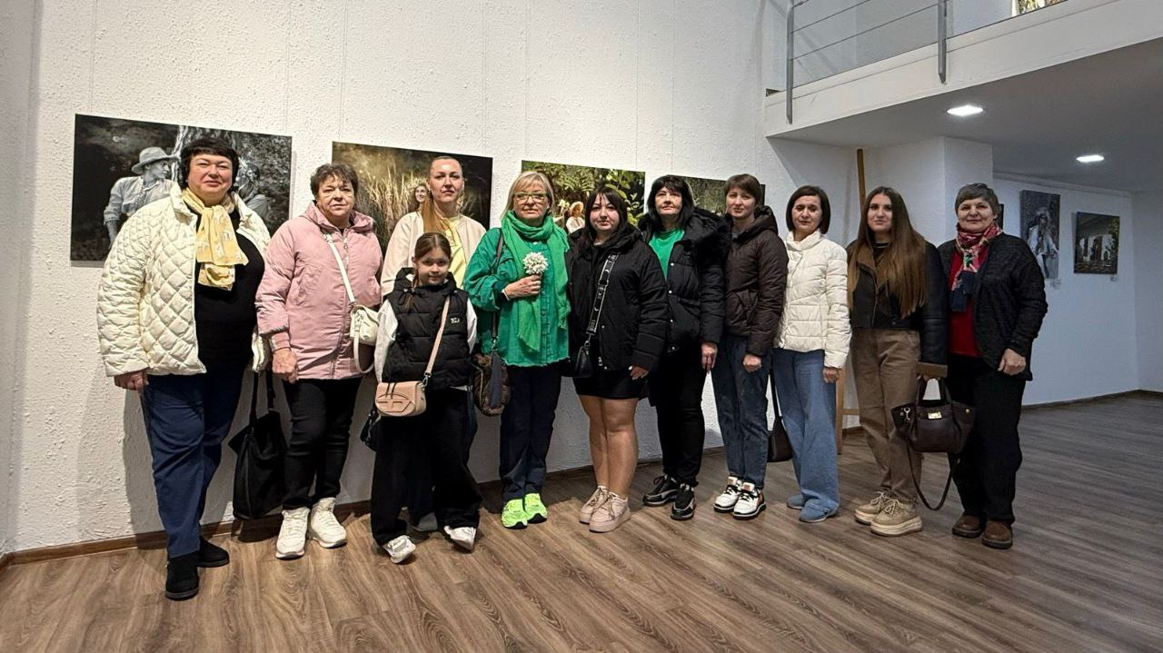 Група жінок та дитина позують для спільного фото в художній галереї на фоні виставлених на стіні картин. Вони одягнені у повсякденний верхній одяг: куртки, пальта та светри. Приміщення має світлі стіни, дерев'яну підлогу та сучасне освітлення.