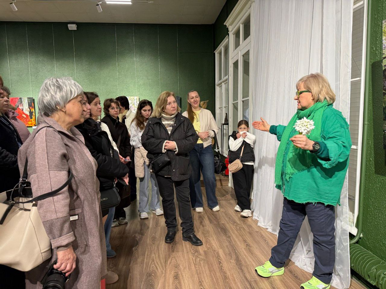 Група людей у художній галереї слухає жінку в яскраво-зеленому вбранні, яка тримає маленьку білу квітку та щось розповідає. Відвідувачі в повсякденному верхньому одязі стоять на дерев'яній підлозі на фоні темно-зелених стін із картинами.