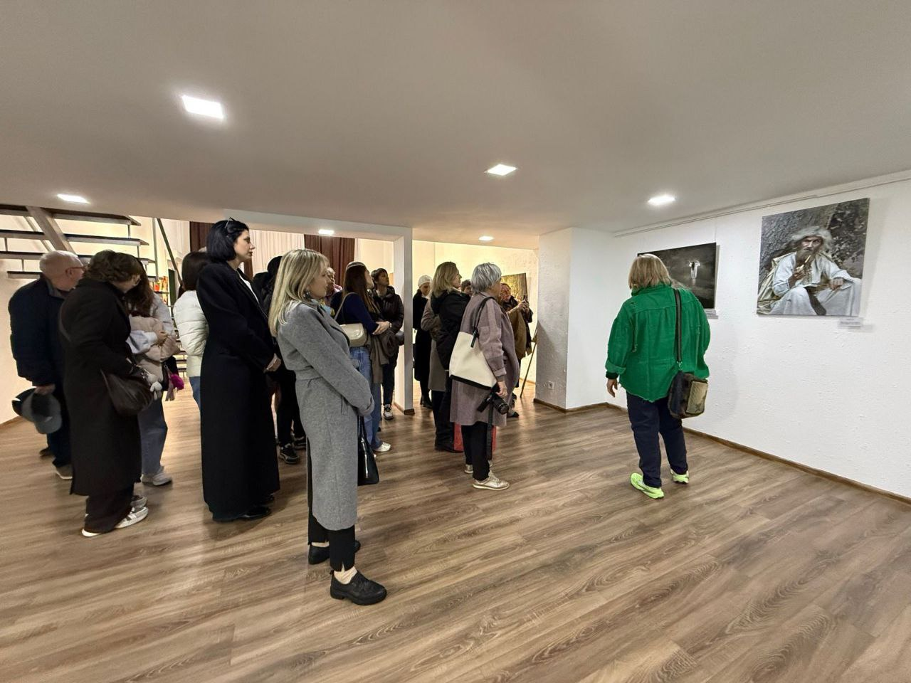 Група відвідувачів у художній галереї розглядає картини на білих стінах. Люди одягнені в демісезонний одяг — пальта, куртки та светри. Приміщення має світле точкове освітлення, дерев'яну підлогу та сходи на задньому плані.