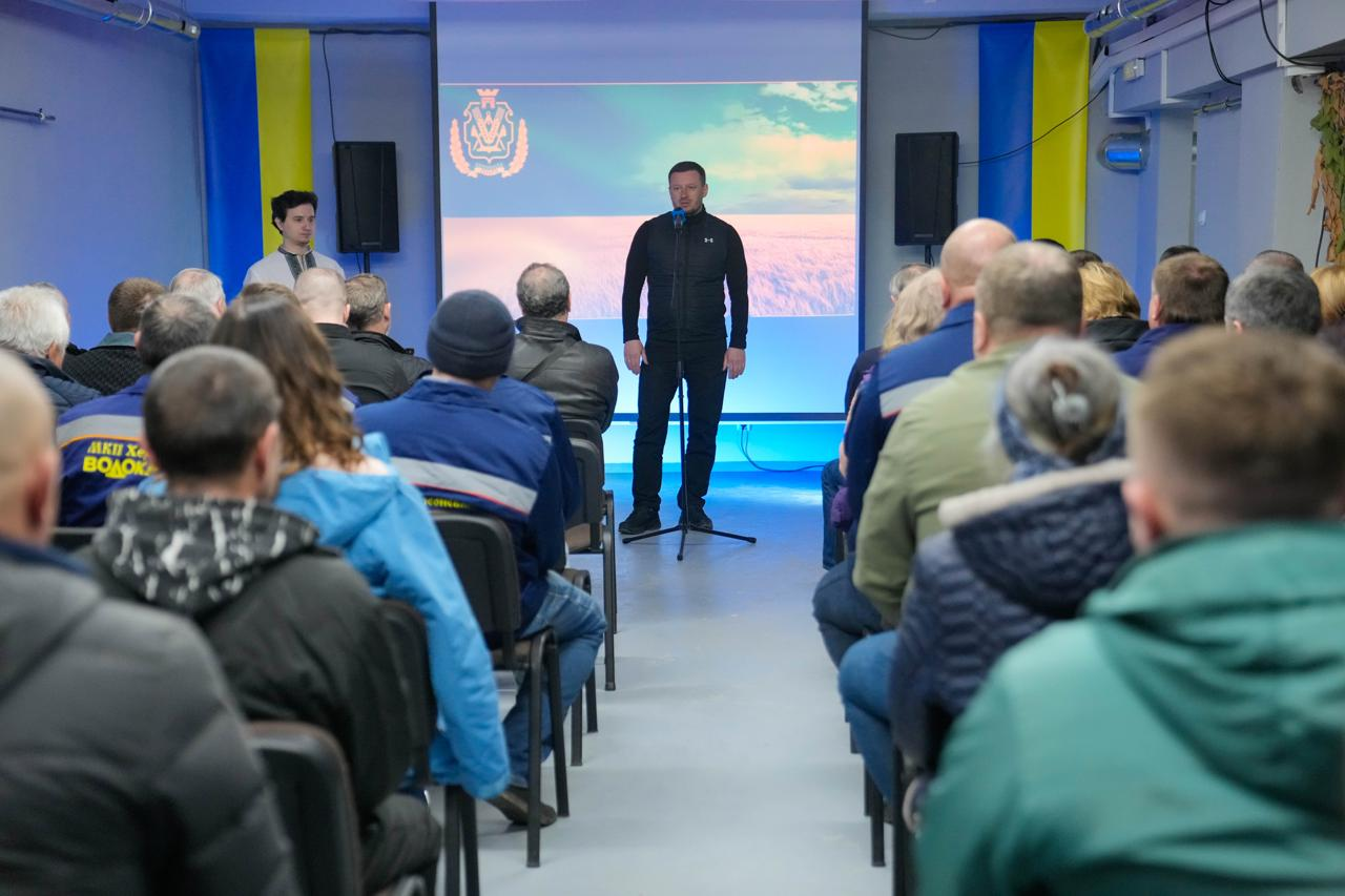 Чоловік виступає перед аудиторією у залі, оформленій у національних кольорах. Позаду нього — екран із зображенням прапора України та герба, а з боків встановлені синьо-жовті стяги. Слухачі сидять спиною до камери, дехто з них одягнений у спецодяг комунальних служб.