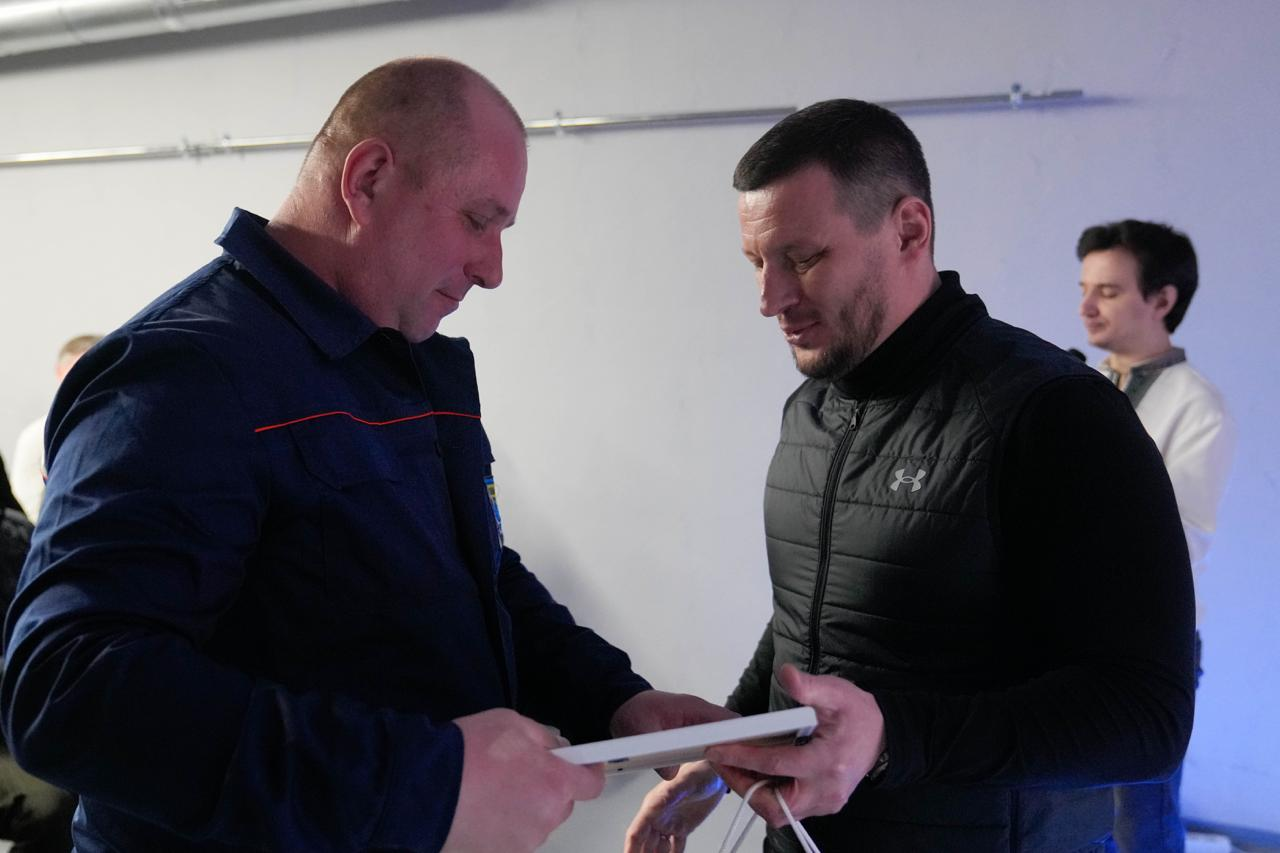 Чоловік у чорному жилеті передає рамку з документом іншому чоловікові в темно-синій робочій формі. Обидва зосереджені на процесі нагородження, на фоні стоїть третя особа. Подія відбувається у світлому приміщенні з мінімалістичним інтер’єром.