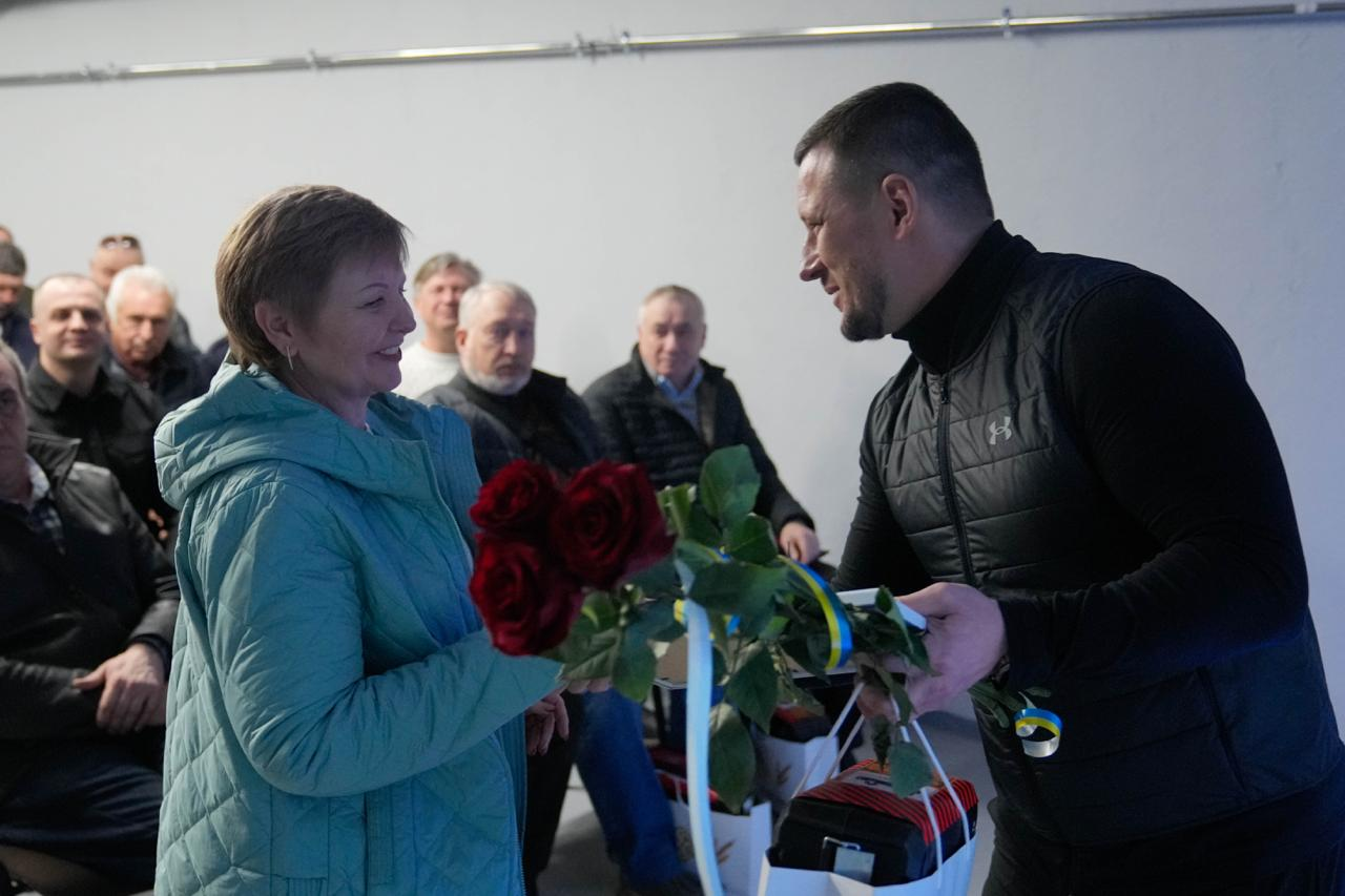 Чоловік у чорному жилеті вручає жінці у блакитній куртці букет червоних троянд, прикрашений синьо-жовтою стрічкою. Обоє посміхаються одне одному під час церемонії. На задньому плані сидять люди, які спостерігають за подією у світлому приміщенні.