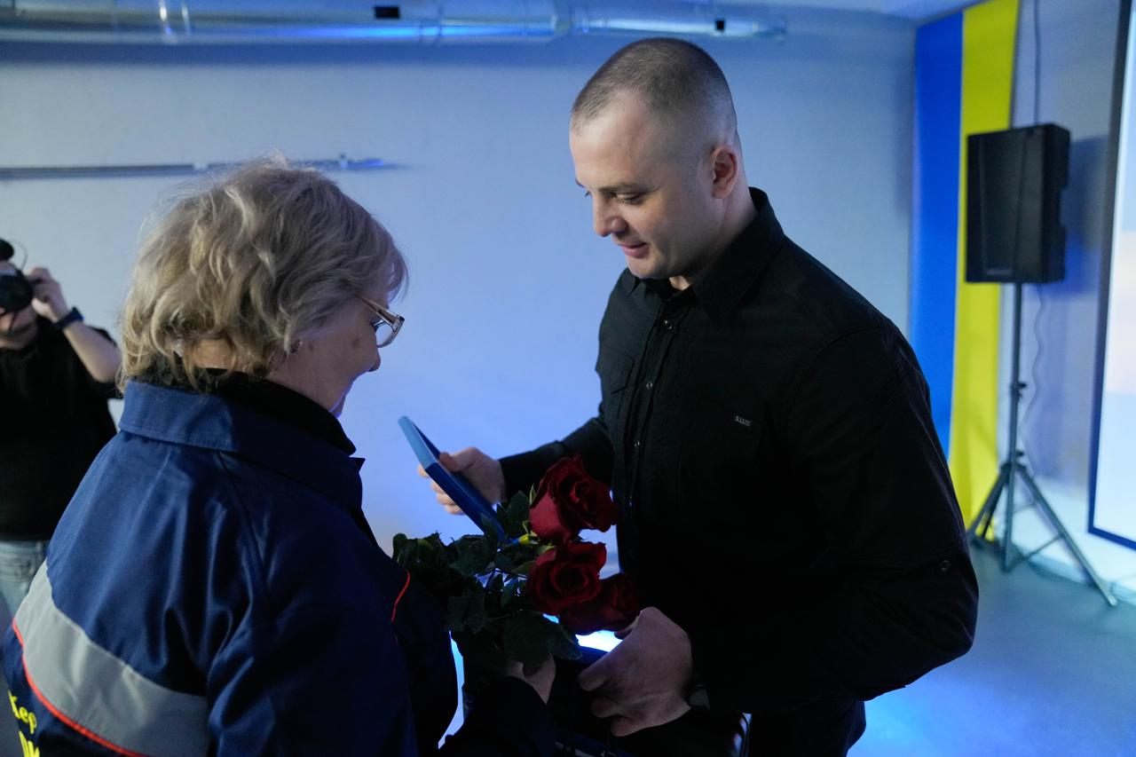 Чоловік у чорній сорочці вручає жінці в окулярах та робочій куртці букет червоних троянд і рамку з нагородою. Позаду них видно синьо-жовтий стяг та звукове обладнання, що вказує на офіційну церемонію в приміщенні.