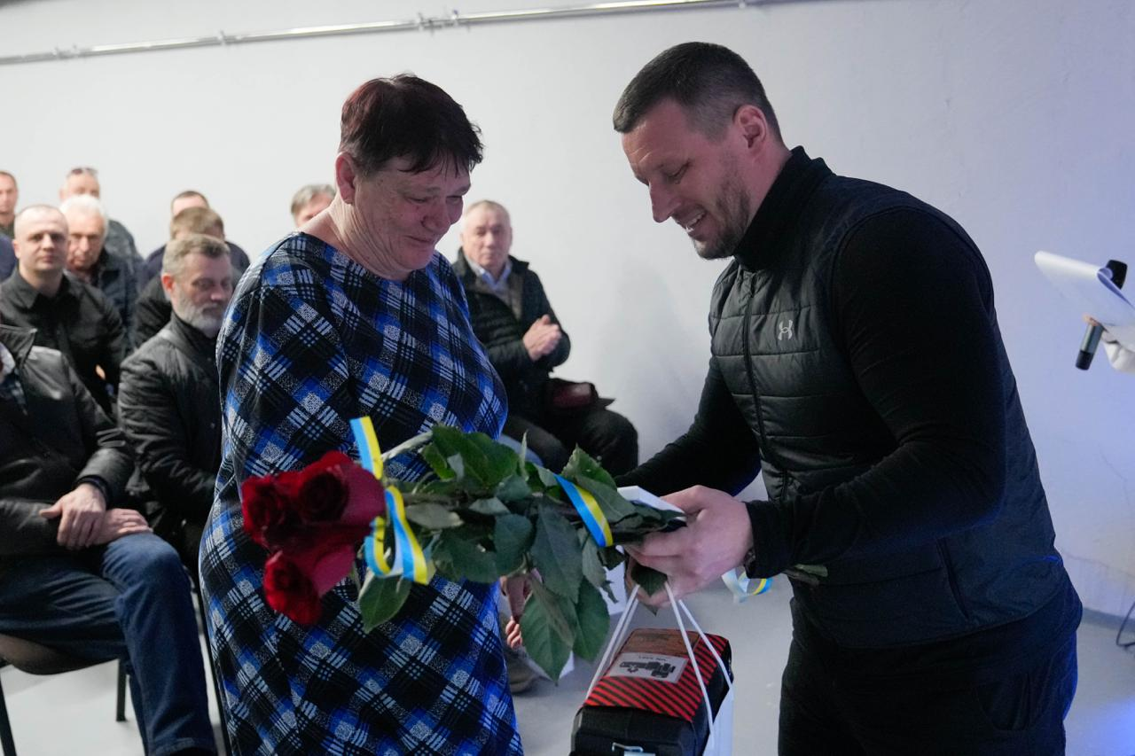 Чоловік у чорному жилеті вручає жінці у картатій сукні букет червоних троянд із синьо-жовтими стрічками та пакунок. Обоє усміхаються під час моменту нагородження. На задньому плані в залі сидять люди, які спостерігають за подією.