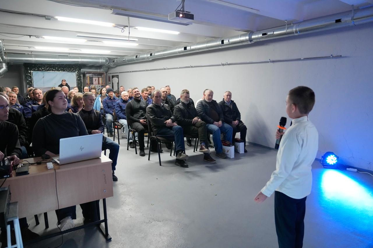 Хлопчик у білій сорочці виступає з мікрофоном перед аудиторією у просторому залі з промисловим дизайном. Слухачі, серед яких чоловіки та жінки, уважно дивляться на дитину. Збоку за столом сидить жінка з ноутбуком, яка забезпечує технічний супровід виступу, а сценічне світло підсвічує підлогу синім кольором.