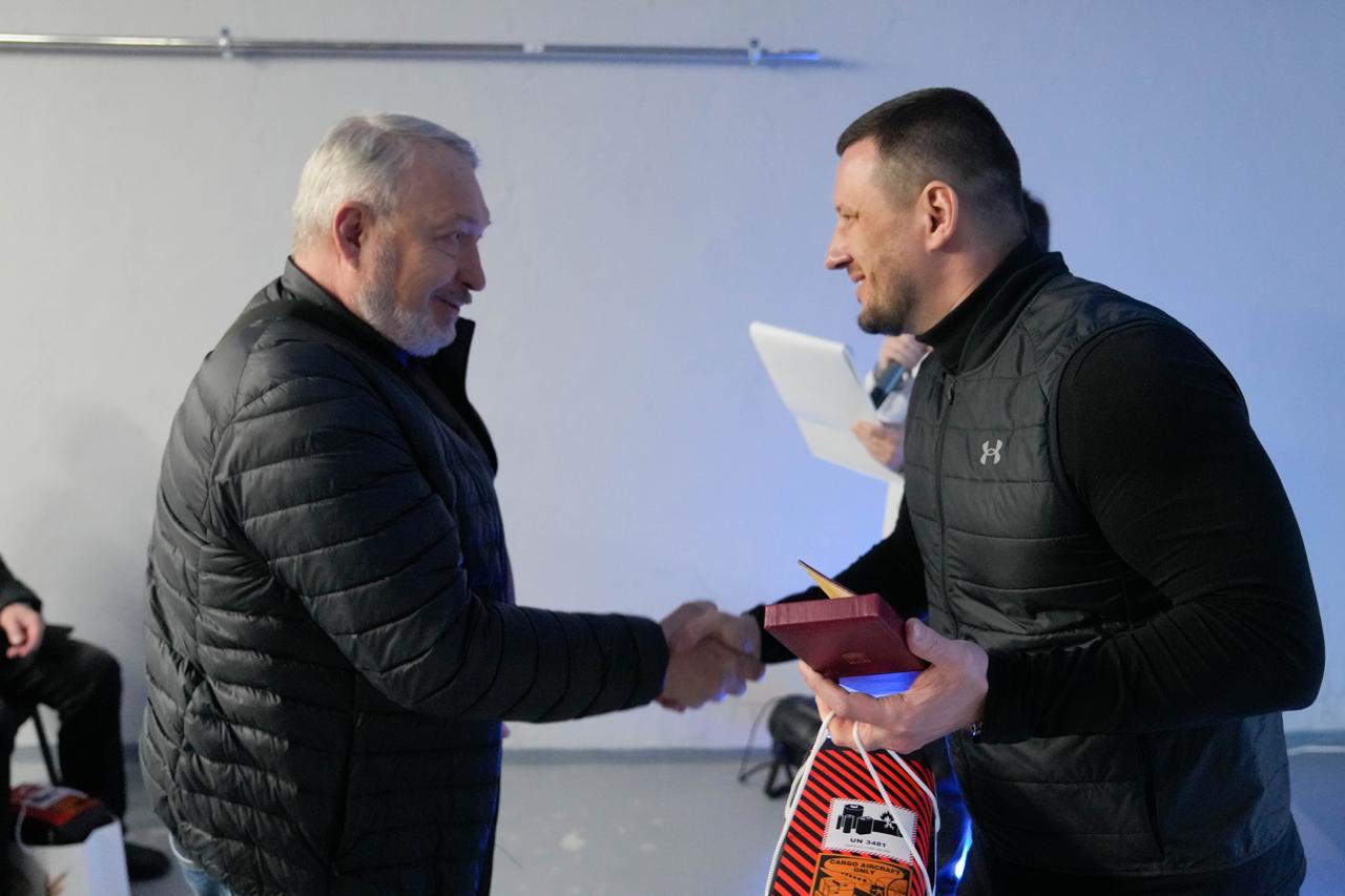 Чоловік у чорному жилеті тисне руку сивочолому чоловікові в куртці, вручаючи йому футляр із нагородою та пакунок. Обидва учасники усміхаються один одному під час моменту нагородження. На задньому плані видно іншу людину з паперами у світлому приміщенні.