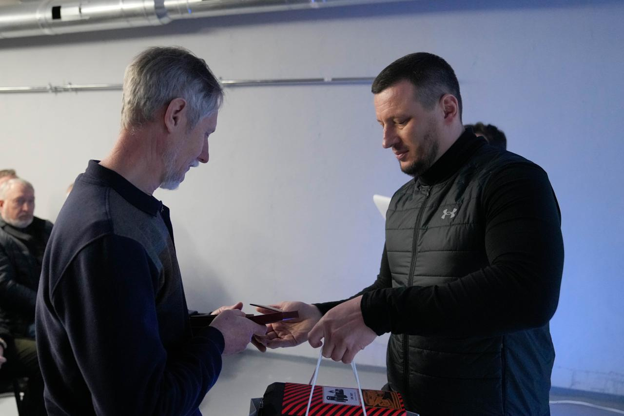 На фото чоловік у чорному жилеті передає іншому чоловікові з сивим волоссям футляр із нагородою та паперовий пакунок. Обидва учасники зосереджені на моменті вручення. Подія відбувається у світлому приміщенні, де на задньому плані видно іншого сивочолого чоловіка, який спостерігає за процесом.