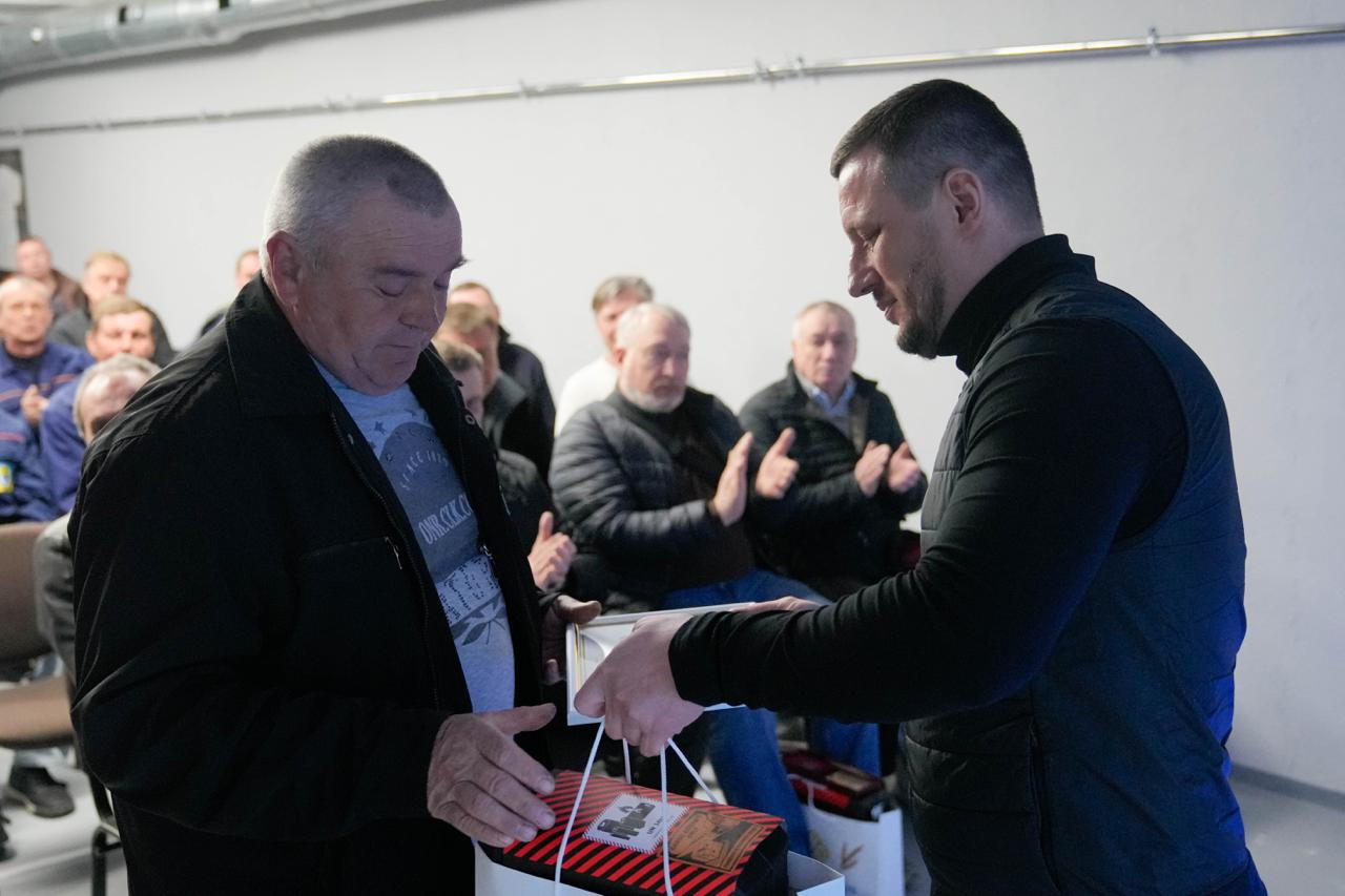 Чоловік у чорному жилеті вручає рамку з документом та паперовий пакунок літньому чоловікові в темній куртці. Обидва зосереджені на моменті нагородження. На задньому плані в залі сидять люди та аплодують, спостерігаючи за подією.