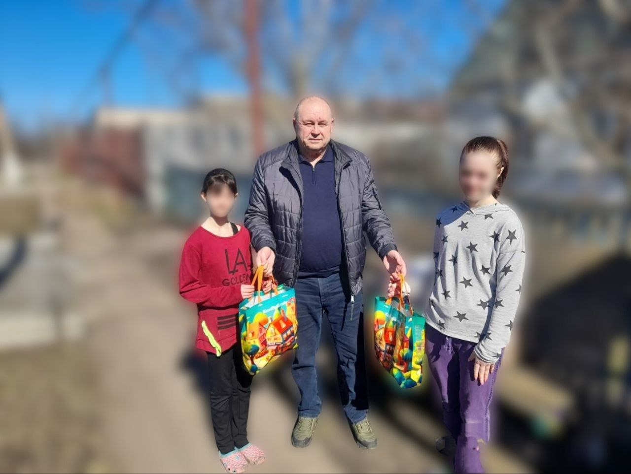 Чоловік у куртці стоїть на вулиці поруч із двома дівчатками та тримає у руках яскраві подарункові пакети з малюнками. Дівчатка стоять по обидва боки від нього, одна в червоному светрі, інша — у сірому з зірочками. Фон зображення розмитий, що підкреслює головних героїв на сонячній вулиці.