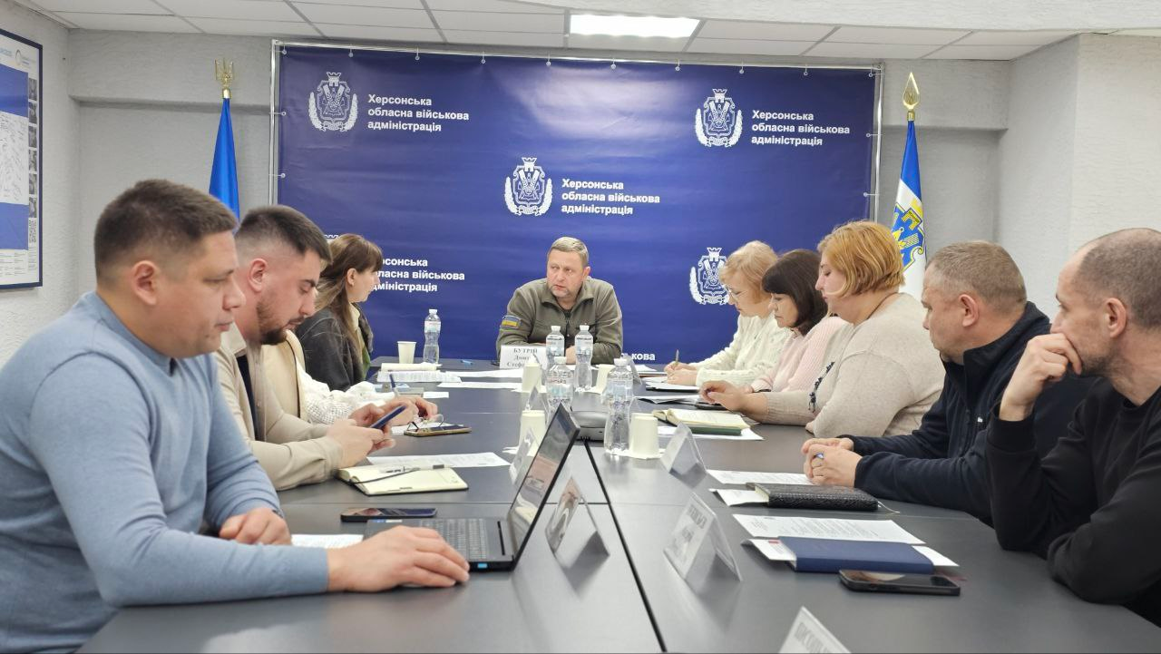 Група людей на робочій нараді за довгим столом. У центрі — чоловік у військовій формі на фоні синього банера з офіційною символікою та державних прапорів. Учасники зустрічі працюють із ноутбуками та роблять записи.