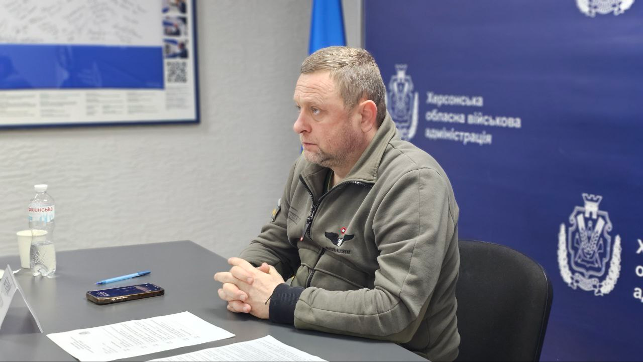 Чоловік у військовій формі сидить за столом під час наради на фоні синього банера з офіційними логотипами. Його руки складені в замок, а перед ним лежать документи, смартфон і стоїть пляшка води. Він зосереджено дивиться вбік, слухаючи співрозмовника поза кадром.