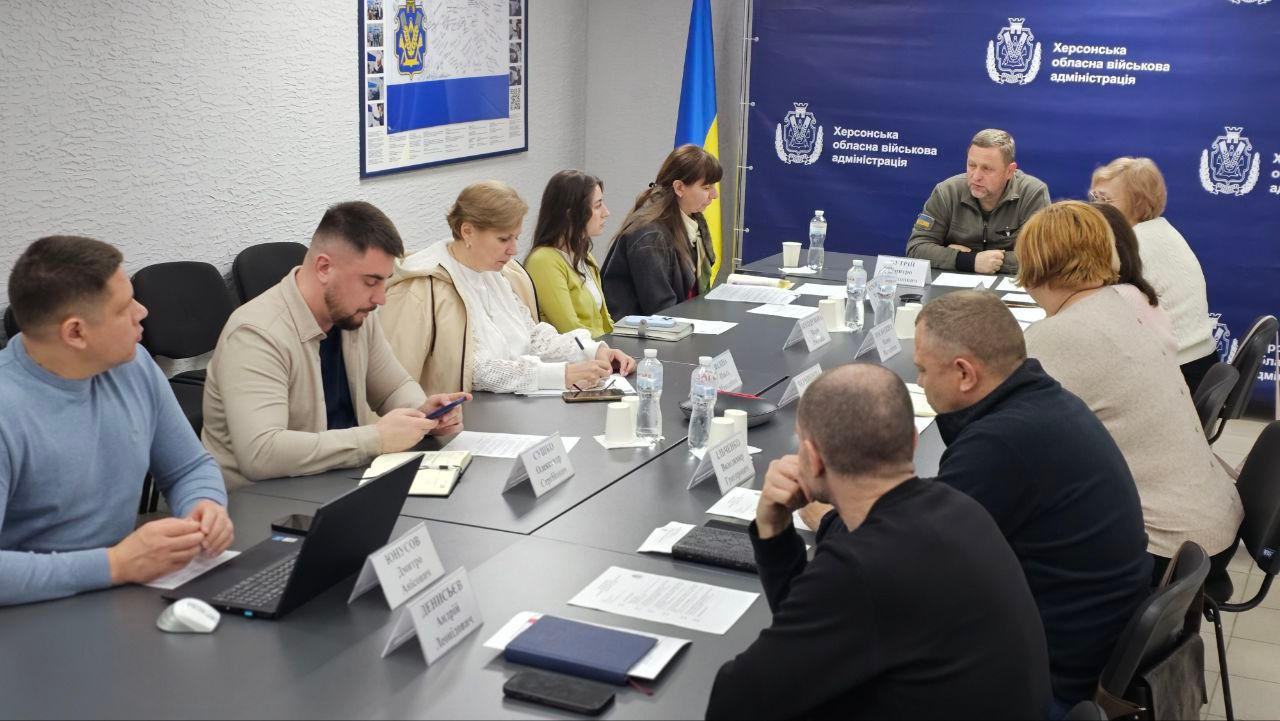 Учасники робочої наради сидять за довгим столом у світлому кабінеті. У центрі — чоловік у військовій формі, який веде обговорення на фоні синього банера з офіційною символікою та державного прапора. Перед присутніми лежать документи, ноутбуки та блокноти для записів.