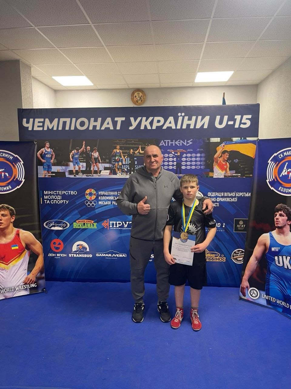 Чоловік у спортивному костюмі та юний борець із медаллю і грамотою стоять на фоні банера «Чемпіонат України U-15». Позаду них видно логотипи спортивних організацій та кадри зі змагань.