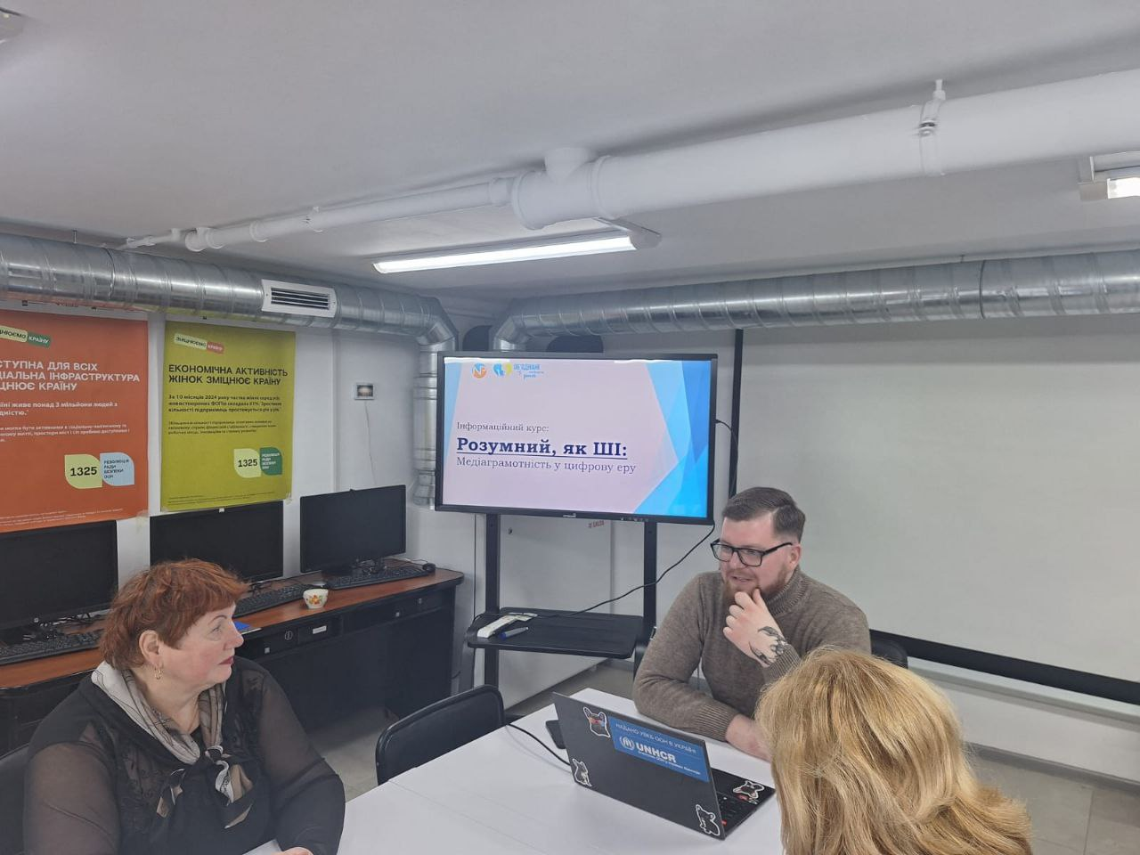Навчальний захід у сучасному офісі: група людей проводить обговорення за столом. На екрані телевізора заголовок інформаційного курсу «Розумний, як ШІ: Медіаграмотність у цифрову еру». На стінах розміщено соціальні плакати, а на ноутбуці видно логотип УВКБ ООН (UNHCR).