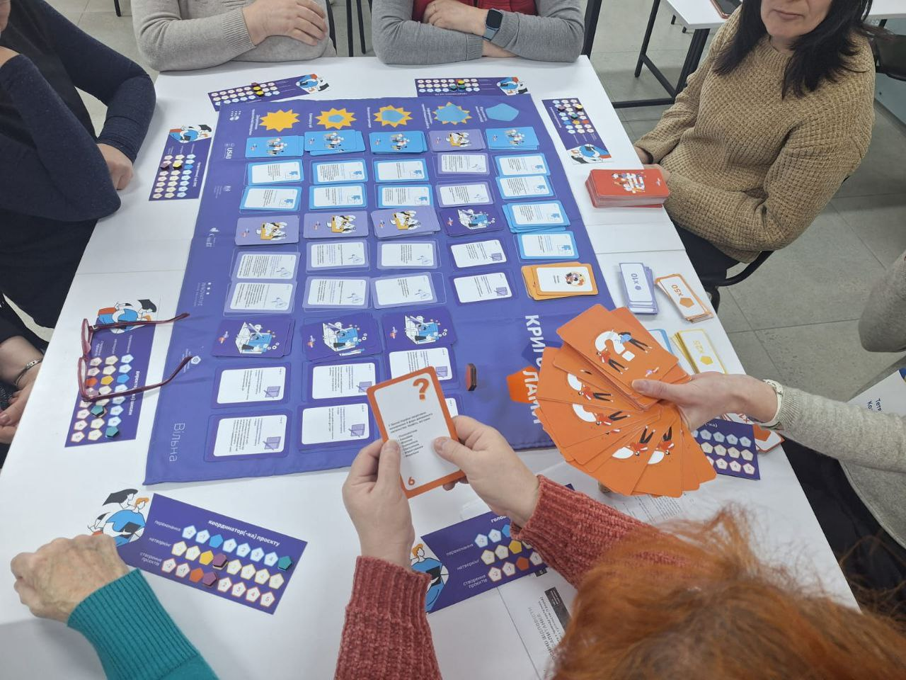 Група людей грає в настільну освітню гру за столом. На великому синьому полі розміщені картки з текстом та іконками, а учасники тримають у руках помаранчеві картки із запитаннями. На полі видно напис «Критичне мислення».