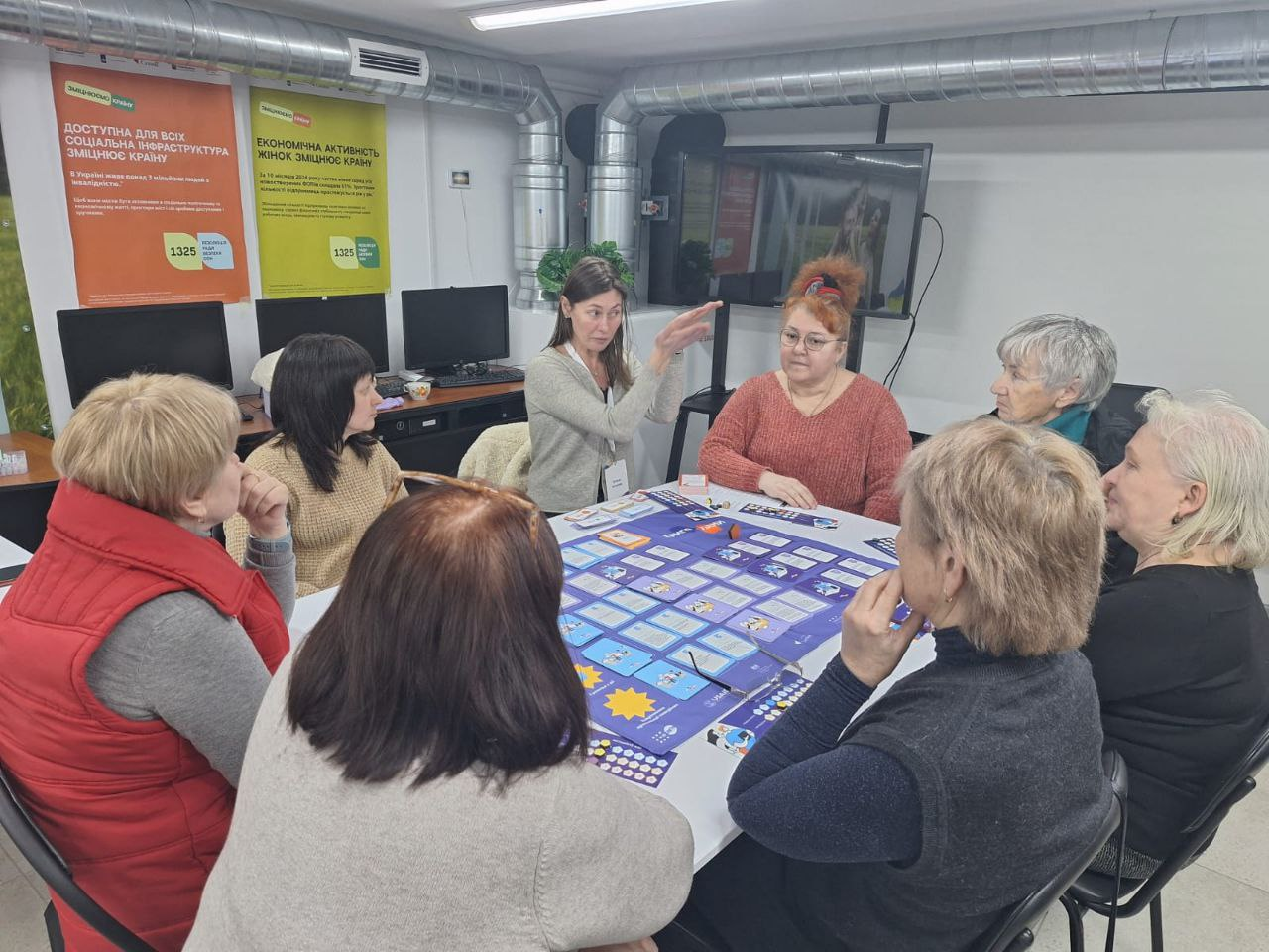 Група жінок проводить навчальне заняття за допомогою великої настільної гри. Учасниці сидять навколо столу з розкладеними картками та ігровим полем. На стіні позаду висять інформаційні плакати про соціальну інфраструктуру та економічну активність жінок.