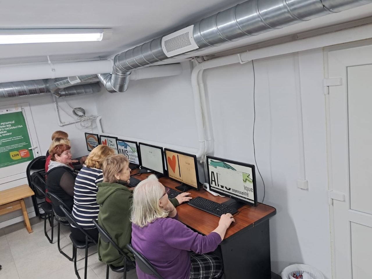 Група жінок займається у комп’ютерному класі, розташованому в облаштованому приміщенні з вентиляційними трубами під стелею. Учасниці сидять за довгим столом, на моніторах якого відображені навчальні матеріали про штучний інтелект.