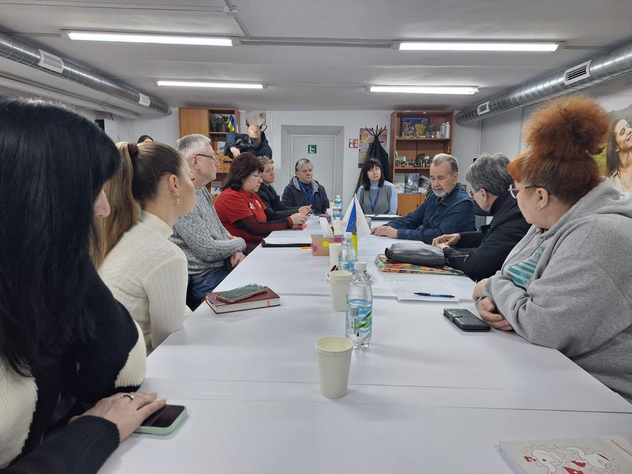 Робоча зустріч або обговорення в офісному приміщенні: група людей сидить за довгим білим столом. На столі розміщено пляшки з водою, смартфони та папери для записів. Приміщення має низьку стелю з відкритими елементами вентиляції.