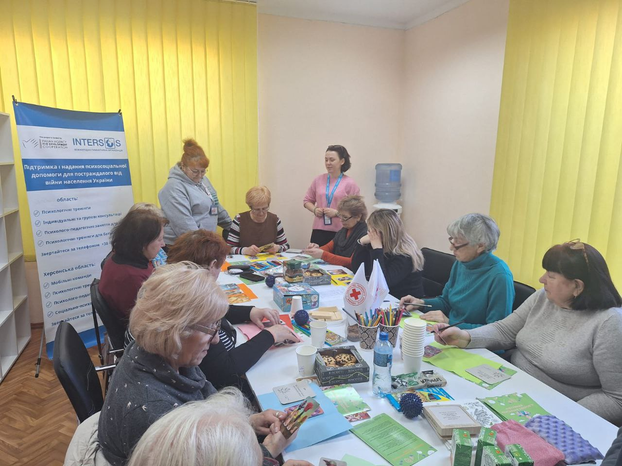 Група жінок займається арттерапією за великим столом у приміщенні з жовтими жалюзі. На столі розкладені олівці, картки, стаканчики та смаколики. Поруч стоїть банер організації INTERSOS про надання психосоціальної допомоги постраждалим від війни.