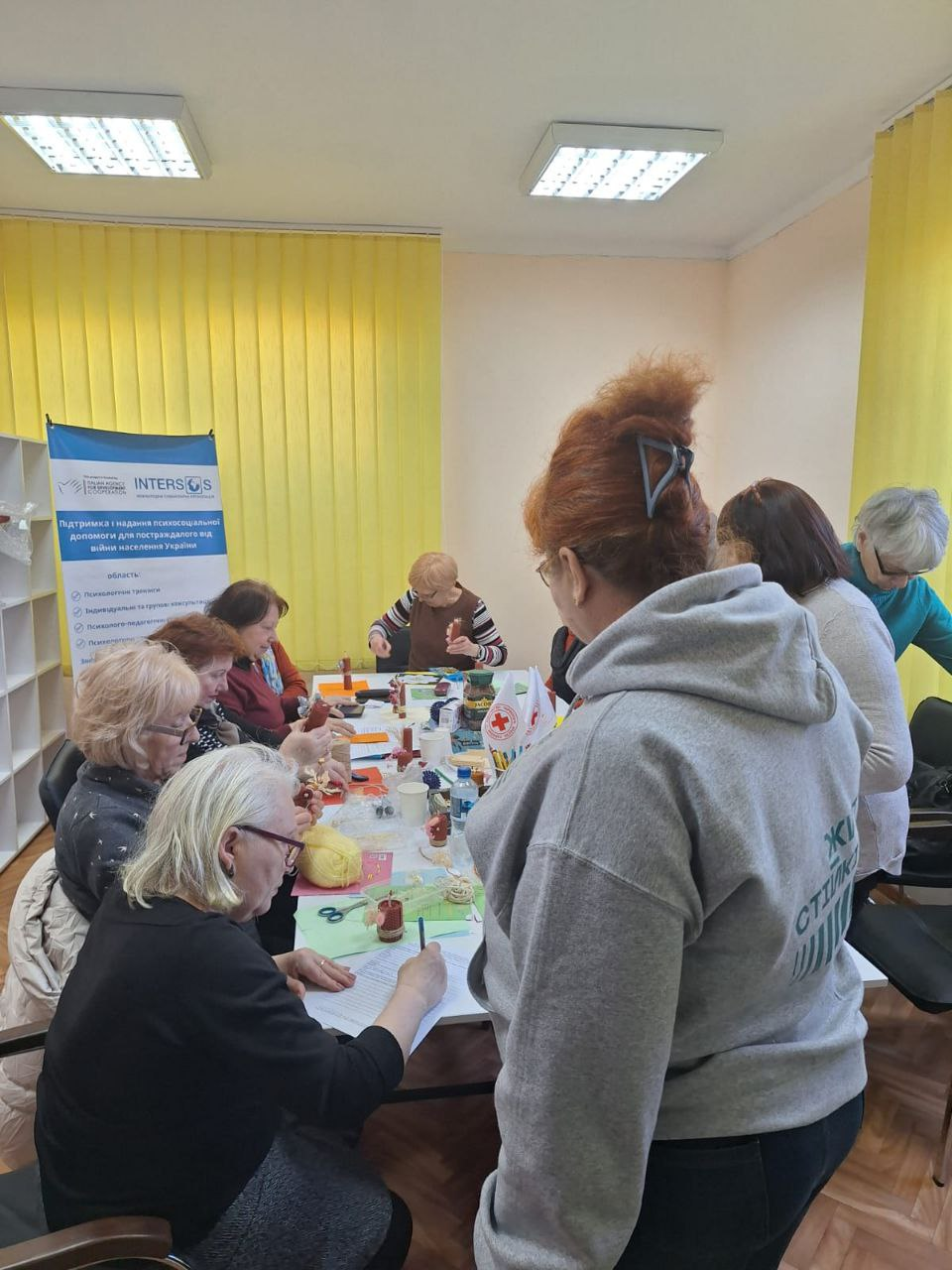 Група жінок займається рукоділлям або арттерапією за великим столом у світлій кімнаті. На столі розкладені матеріали для творчості та прапорці Товариства Червоного Хреста України. Позаду стоїть банер гуманітарної організації INTERSOS.