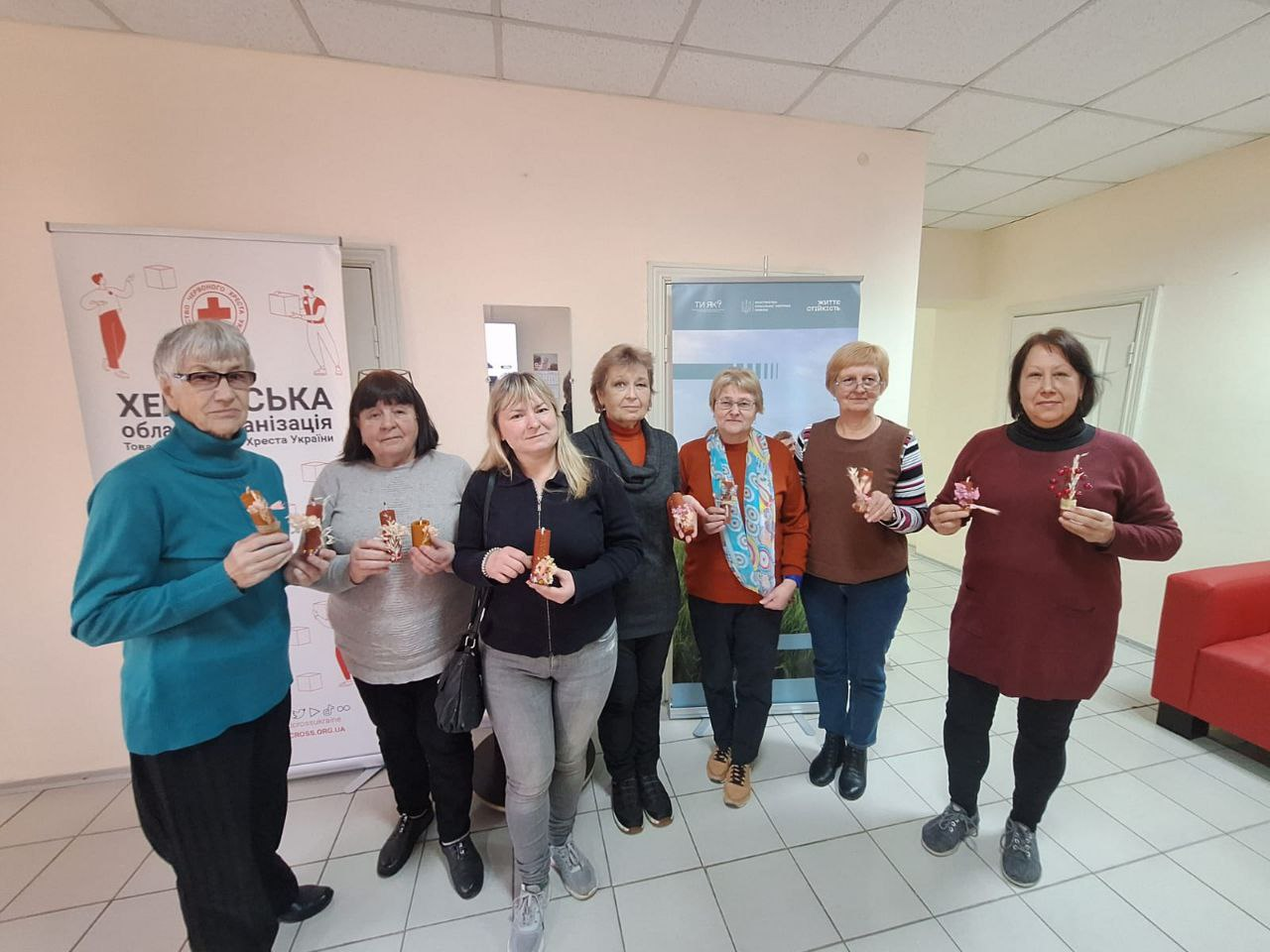 Група жінок демонструє виготовлені обереги у світлому приміщенні. Позаду стоять інформаційні банери Херсонської обласної організації Товариства Червоного Хреста України та гуманітарних партнерів.