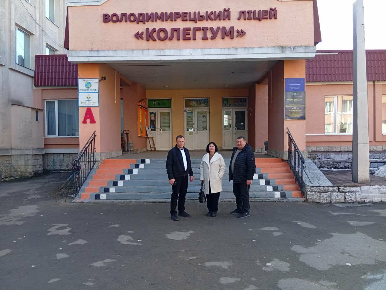 Три людини стоять перед входом до будівлі з написом «Володимирецький ліцей "Колегіум"». Вони позують на фоні широких сходів та центрального входу до навчального закладу.