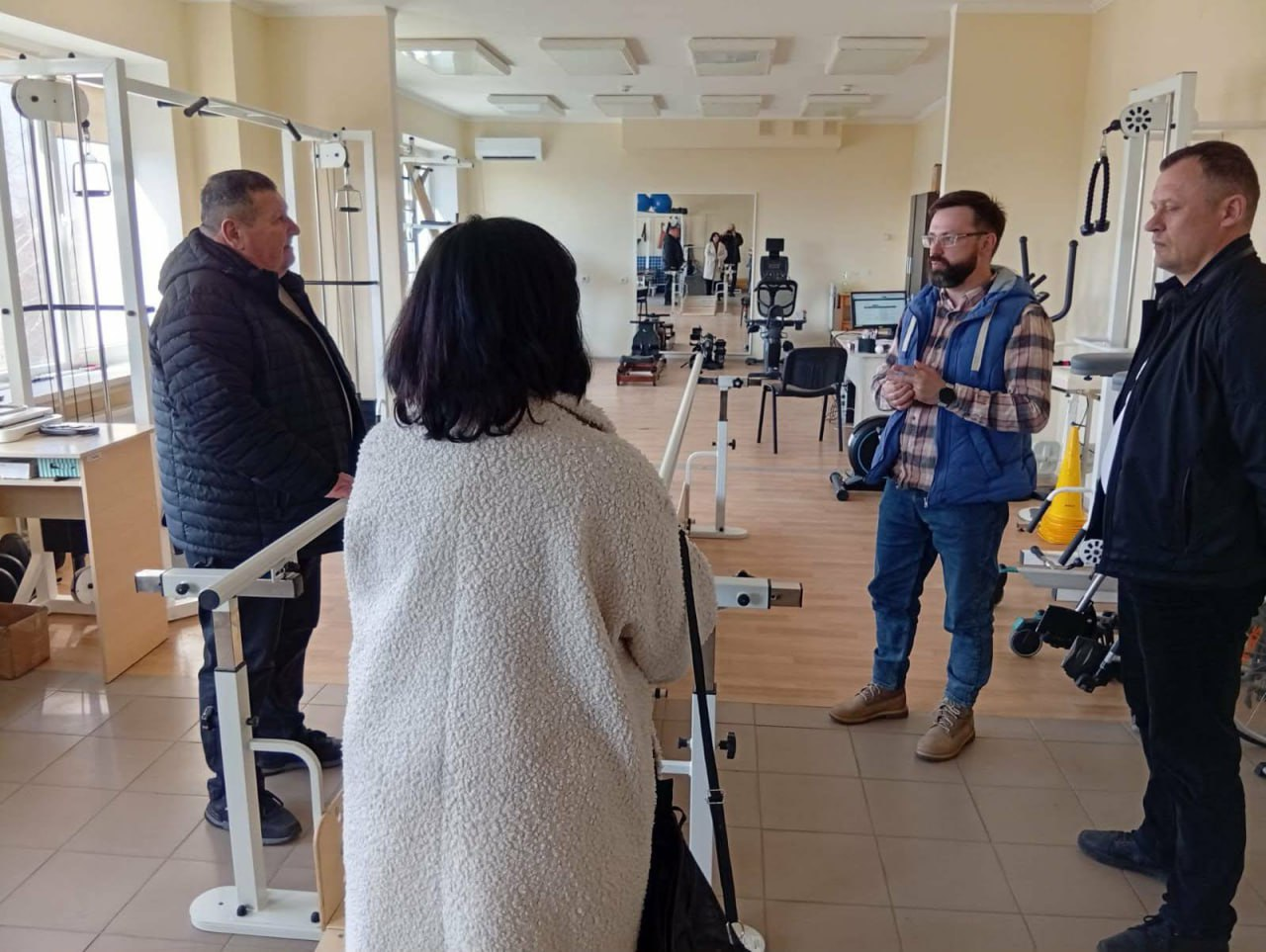 Чотири особи спілкуються у просторому світлому залі для реабілітації, обладнаному різноманітними тренажерами та спеціальними пристроями для фізичних вправ.