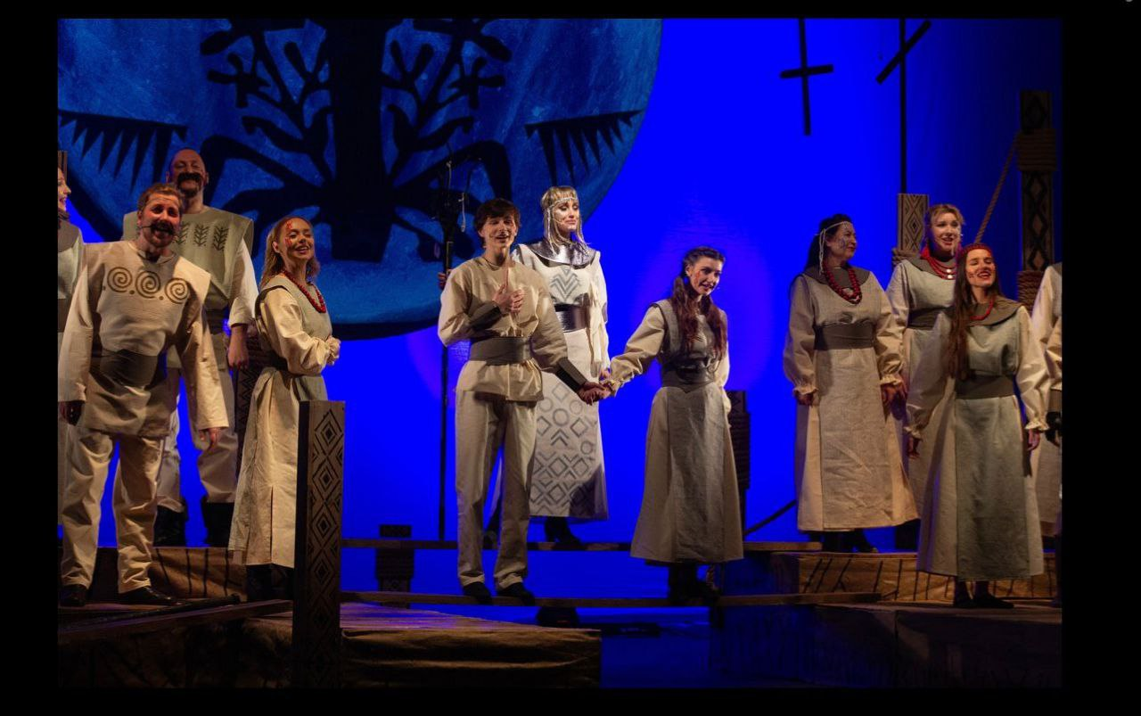Театральна сцена зі співаками в автентичних світлих костюмах з українськими орнаментами. У центрі молода пара тримається за руки, навколо них — хор. На фоні насиченого синього світла видно великий декоративний елемент, що нагадує Дерево життя.