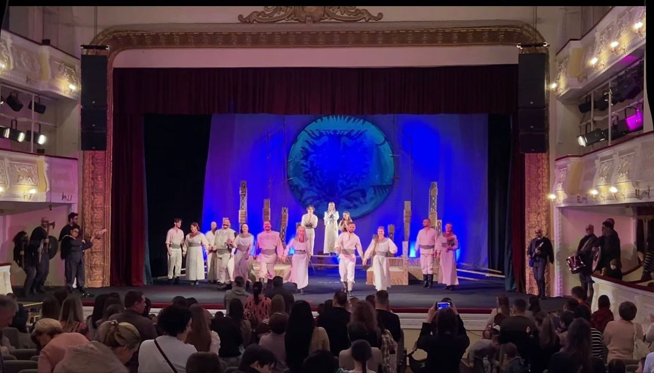 Фінальна сцена театральної вистави: група акторів у світлих стилізованих костюмах виходить на уклін до глядачів. На задньому плані сцени — кругла декорація з символічним орнаментом на синьому фоні. Зал заповнений глядачами, які спостерігають за дійством.