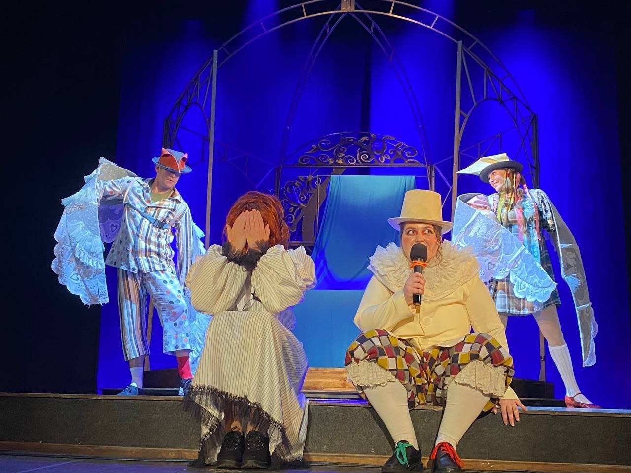 Чотири актори в яскравих театральних костюмах на сцені з синім підсвічуванням. Попереду одна людина закриває обличчя руками, а інша в циліндрі говорить у мікрофон. На задньому плані двоє акторів у капелюхах з великими мереживними крилами стоять біля ажурної металевої арки.