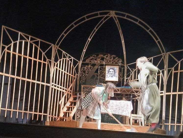 Театральна сцена з дерев’яними ажурними декораціями у формі арок. Двоє акторів у костюмах взаємодіють на тлі столу з білою скатертиною та невеликого портрета, закріпленого на конструкції.