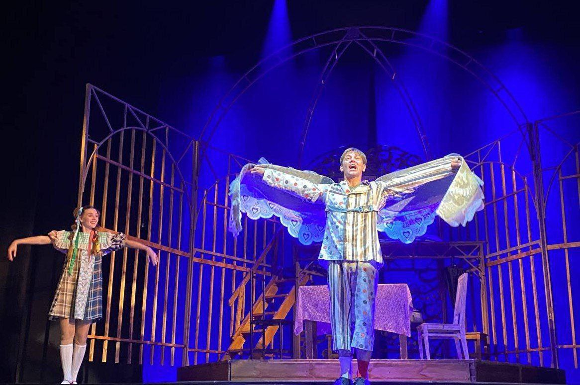 Театральна сцена з двома акторами на синьому фоні під софітами. На передньому плані актор у смугастому костюмі широко розвів руки, тримаючи декоративну накидку з візерунком у формі сердець. Позаду, за металевою ґратчастою конструкцією, стоїть акторка у картатій сукні.