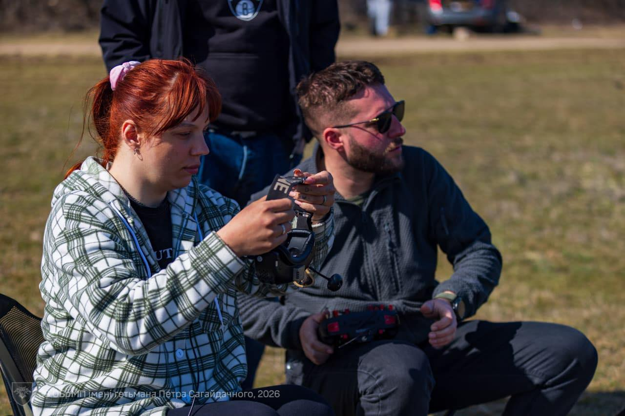 Двоє людей сидять на відкритому просторі та налаштовують обладнання для керування дроном. Жінка в картатій сорочці тримає FPV-окуляри, а чоловік поруч тримає пульт дистанційного керування.