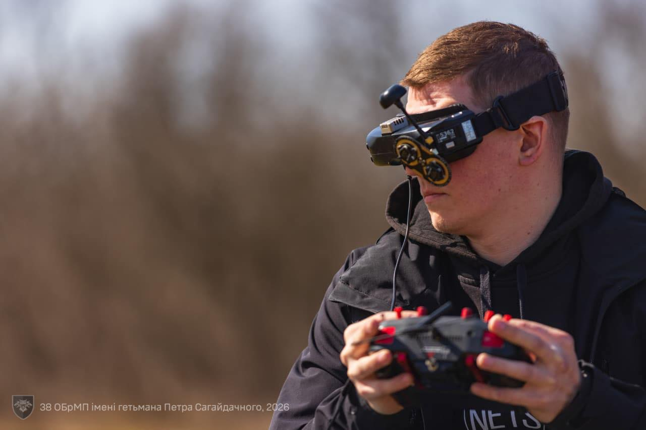 Чоловік у чорному худі керує дроном за допомогою пульта та FPV-окулярів. На фоні видно розмите поле, а в нижньому куті розміщено логотип 38-ї окремої бригади морської піхоти.