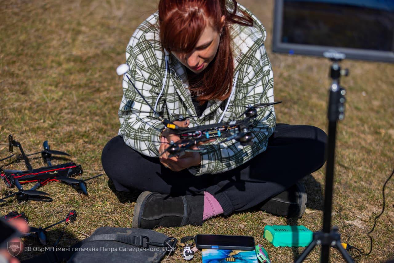 Жінка в картатій сорочці сидить на траві та налаштовує FPV-дрон. Поруч на землі лежить інший квадрокоптер, смартфон та акумулятори. В нижньому куті зображення — логотип 38-ї окремої бригади морської піхоти.