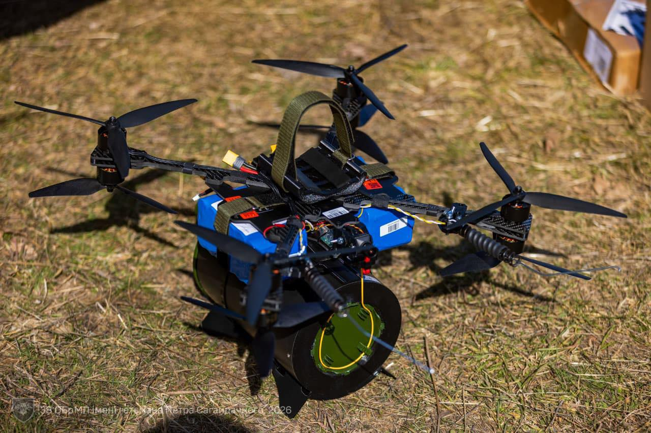 Великий восьмигвинтовий дрон (октокоптер) стоїть на землі. До його корпусу прикріплено два сині акумулятори та габаритний циліндричний предмет знизу. У лівому нижньому куті розміщено водяний знак 38-ї окремої бригади морської піхоти.