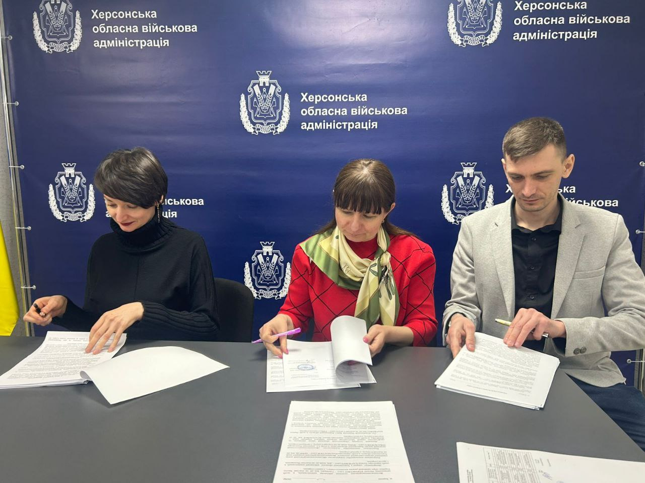 Три особи сидять за столом і підписують офіційні документи на фоні синього брендованого банера з написом «Херсонська обласна військова адміністрація» та гербом області.