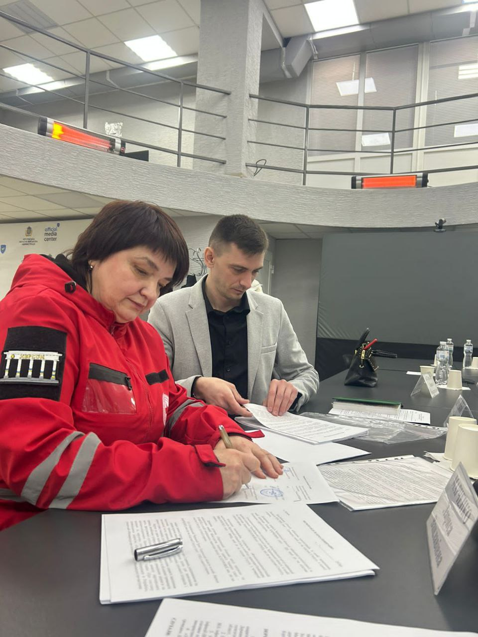 Двоє осіб сидять за столом у сучасному приміщенні та вивчають документи. Жінка в червоній куртці з емблемою Херсона підписує папери, а чоловік поруч переглядає іншу частину документації.