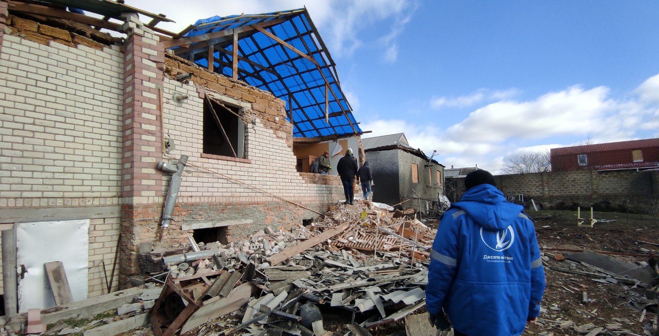 Люди на подвір'ї оглядають значні руйнування житлового будинку: стіни частково обвалені, дах розбитий і накритий синьою плівкою. На передньому плані стоїть людина у синій куртці з логотипом благодійної організації «Десяте квітня» на фоні будівельного сміття.