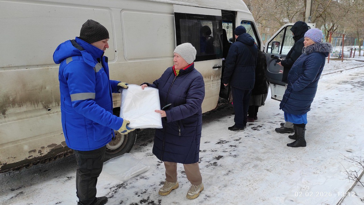 На засніженій вулиці біля білого мікроавтобуса чоловік у синій куртці передає літній жінці пакунок із білим матеріалом. Поруч стоять інші люди, а на зображенні видно дату — 02.02.2026.