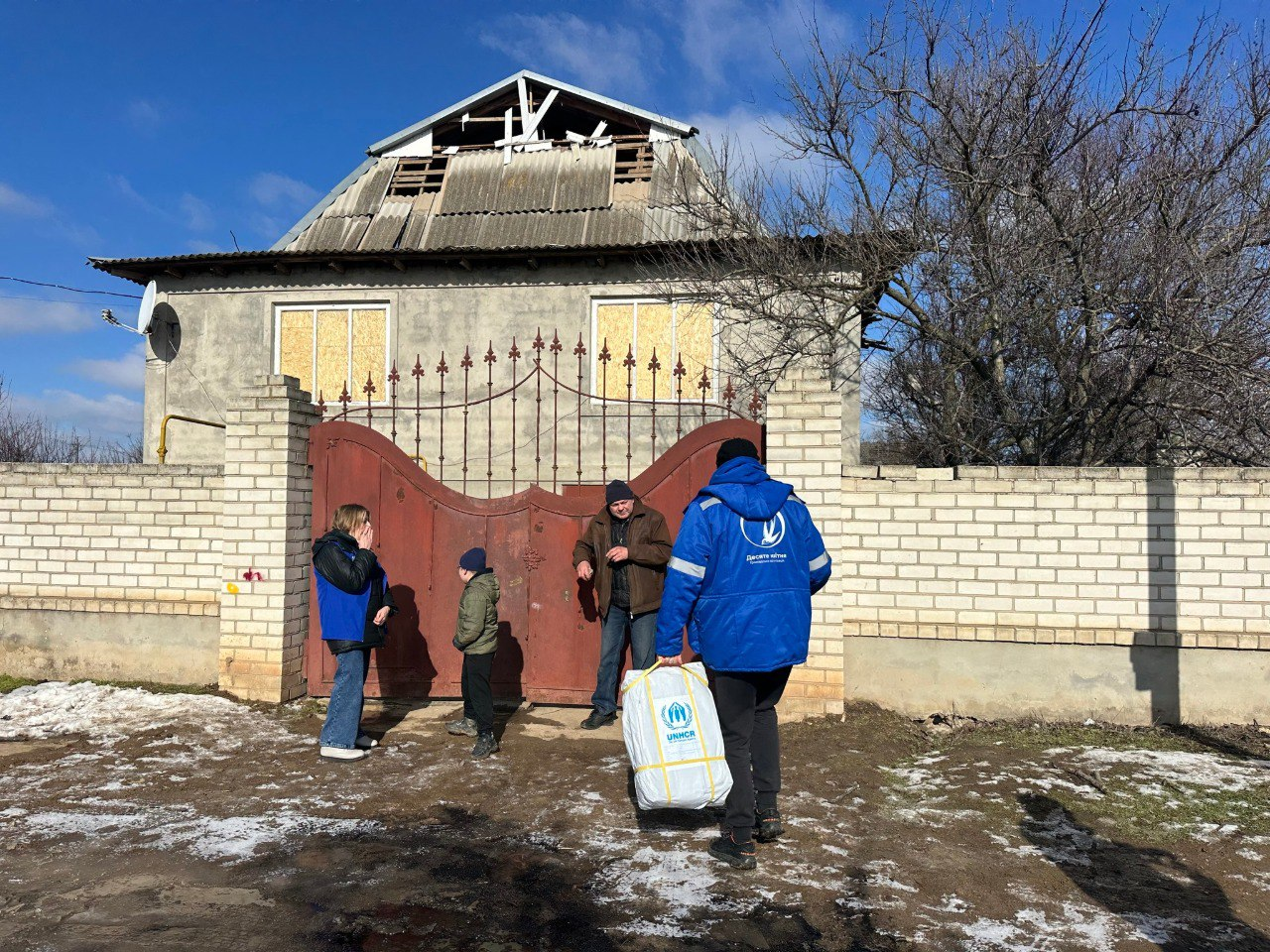Волонтери у синьому одязі з логотипами благодійних організацій привезли гуманітарну допомогу в білому пакунку родині до приватного будинку. На фоні видно пошкоджену обстрілами будівлю із забитими вікнами та зруйнованим дахом.